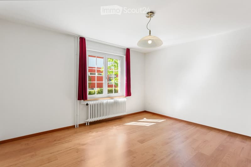 Empty room with wooden flooring, white walls, large window with red curtains, radiator, and a hanging light fixture.
