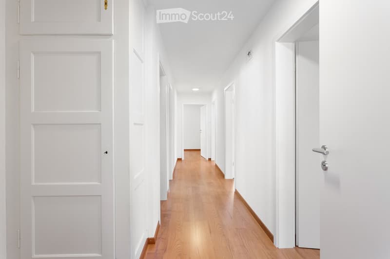 A clean, modern hallway with white walls, wooden flooring, and multiple white doors on both sides.