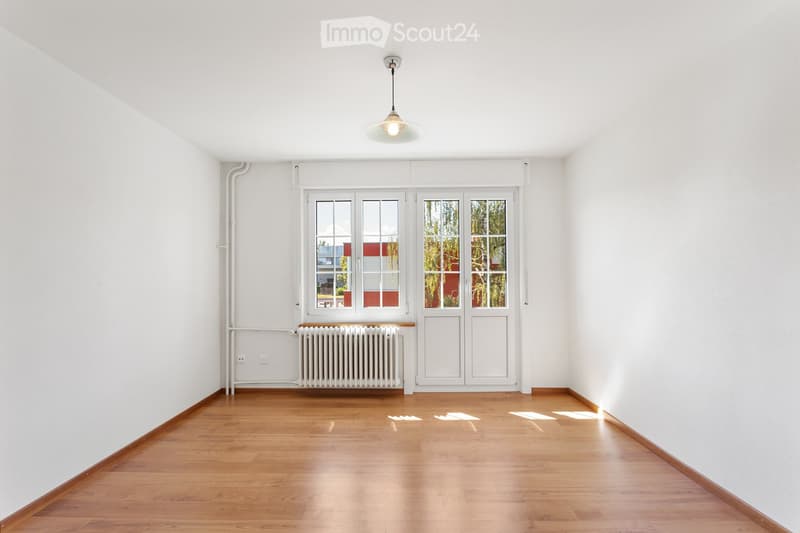 Empty room with white walls, wooden floor, large windows, radiator, and a single hanging light fixture.