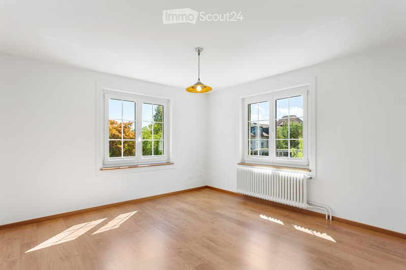Empty room with white walls, wooden flooring, two windows, radiator, and a hanging light fixture.