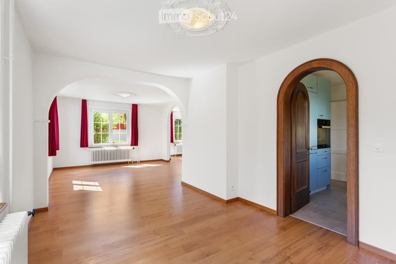 Empty room with wooden flooring, white walls, large windows with red curtains, and a radiator. An arched doorway leads to a kitchen area with blue cabinets and stainless steel appliances.