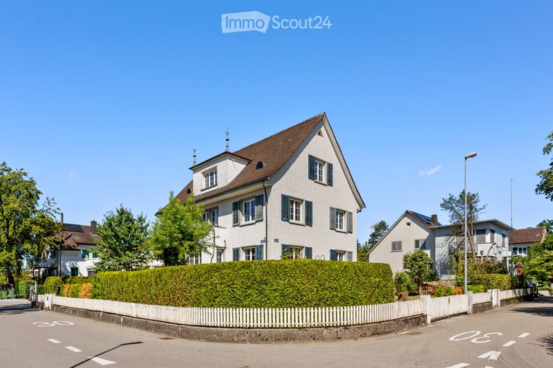 A two-story house with a pitched roof, white exterior, multiple windows, and a well-maintained hedge in front.