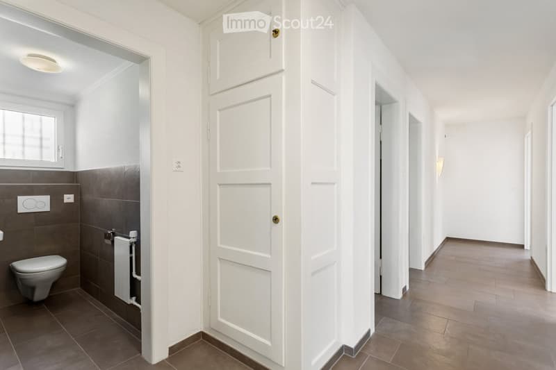 A modern, clean hallway with white walls and tiled flooring, leading to a bathroom with a toilet and dark brown tiles.