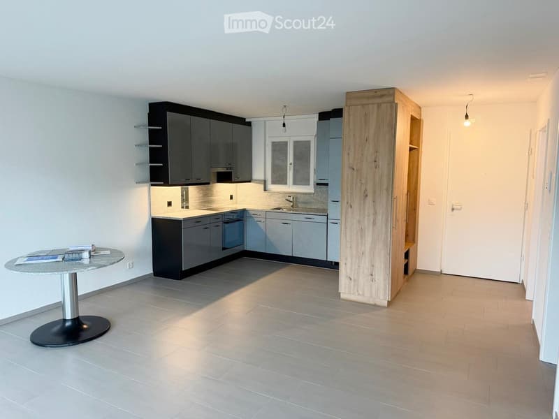 Modern kitchen with grey cabinets, a central island, a round table, and built-in appliances. The space features tiled flooring and a large window.