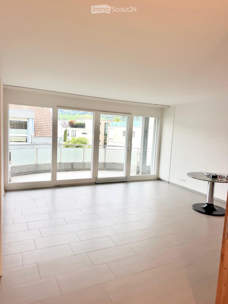Empty room with white tiled flooring, large sliding glass doors leading to a balcony, and a small round table with books in the corner.