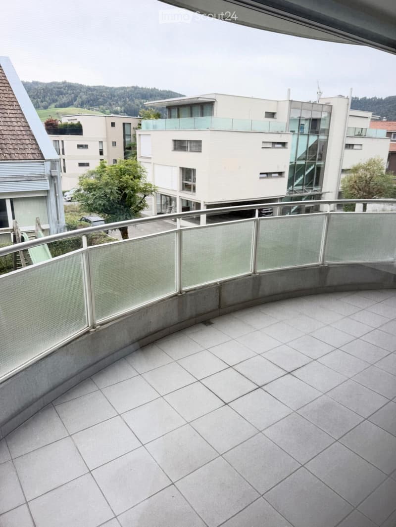 Balcony with tiled floor, glass railing, and a view of surrounding buildings and greenery.