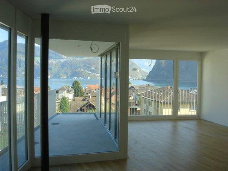 Bright, empty room with large windows and a view of a lake and mountains. Wooden flooring and white walls.