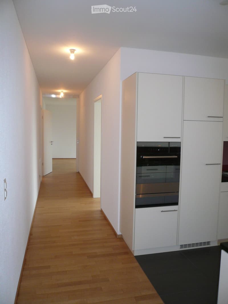 A narrow hallway with light wooden flooring, white walls, and recessed lighting. The hallway leads to a partially visible kitchen area with white cabinets and stainless steel appliances.