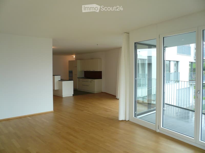 Bright, open living space with wooden flooring, large sliding glass doors leading to a balcony, and a modern kitchen area with white cabinets and a countertop.