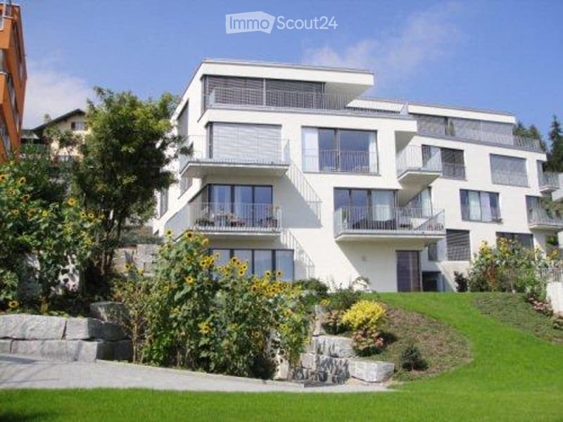 Modern white multi-story apartment building with balconies and large windows, surrounded by a well-maintained garden with sunflowers and shrubs.