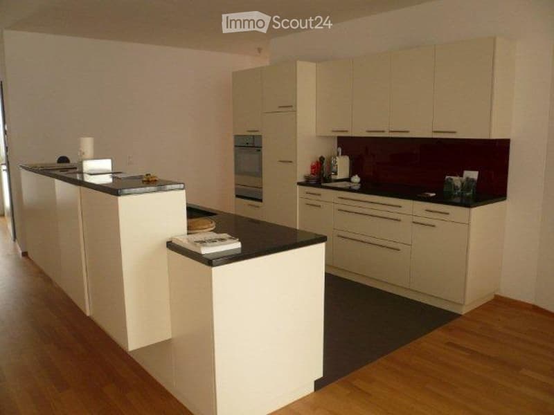 Modern kitchen with light-colored cabinets, black countertops, and a central island. Features include an oven, sink, and various small items on the counters.