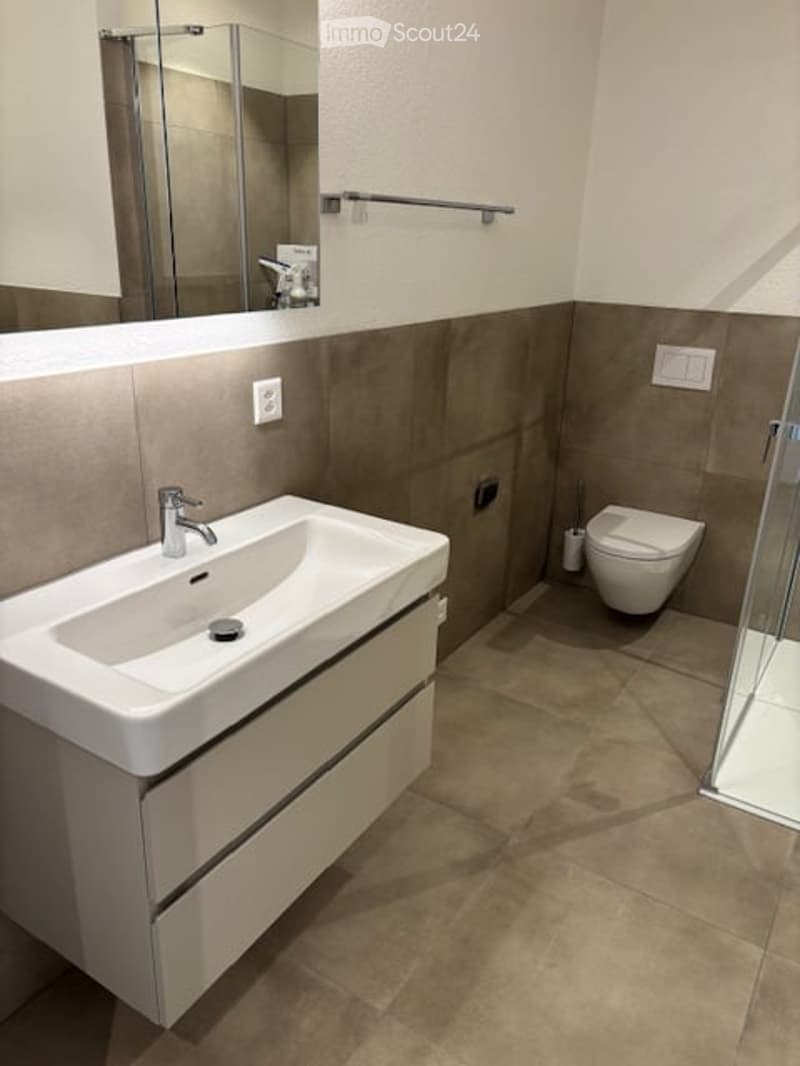 Modern bathroom with a white sink, mirror, toilet, and glass-enclosed shower. Beige tiles and neutral tones.