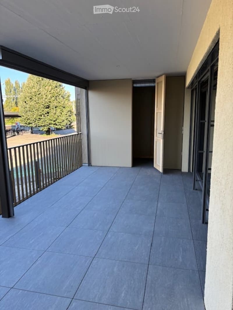 Balcony with grey tiled floor, metal railing, open sliding doors leading to interior, view of greenery and parking area.