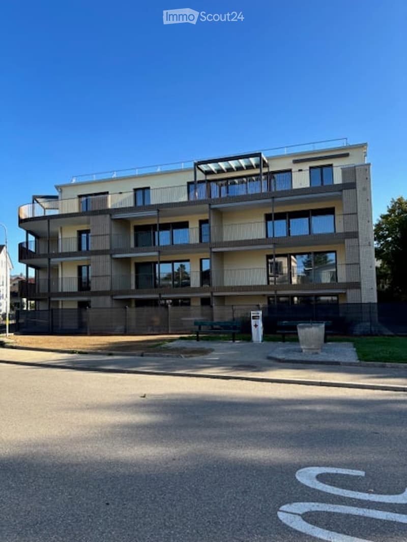 Modern multi-story apartment building with balconies and large windows, surrounded by a small landscaped area with benches.