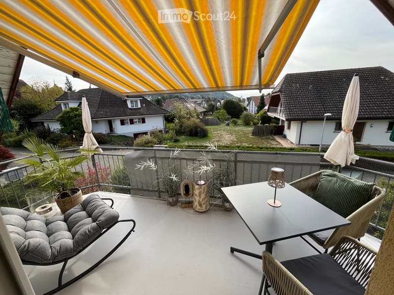 A modern balcony with a grey sofa, a black dining table, wicker chairs, and potted plants, featuring a yellow and white striped awning and a scenic view of the neighborhood.