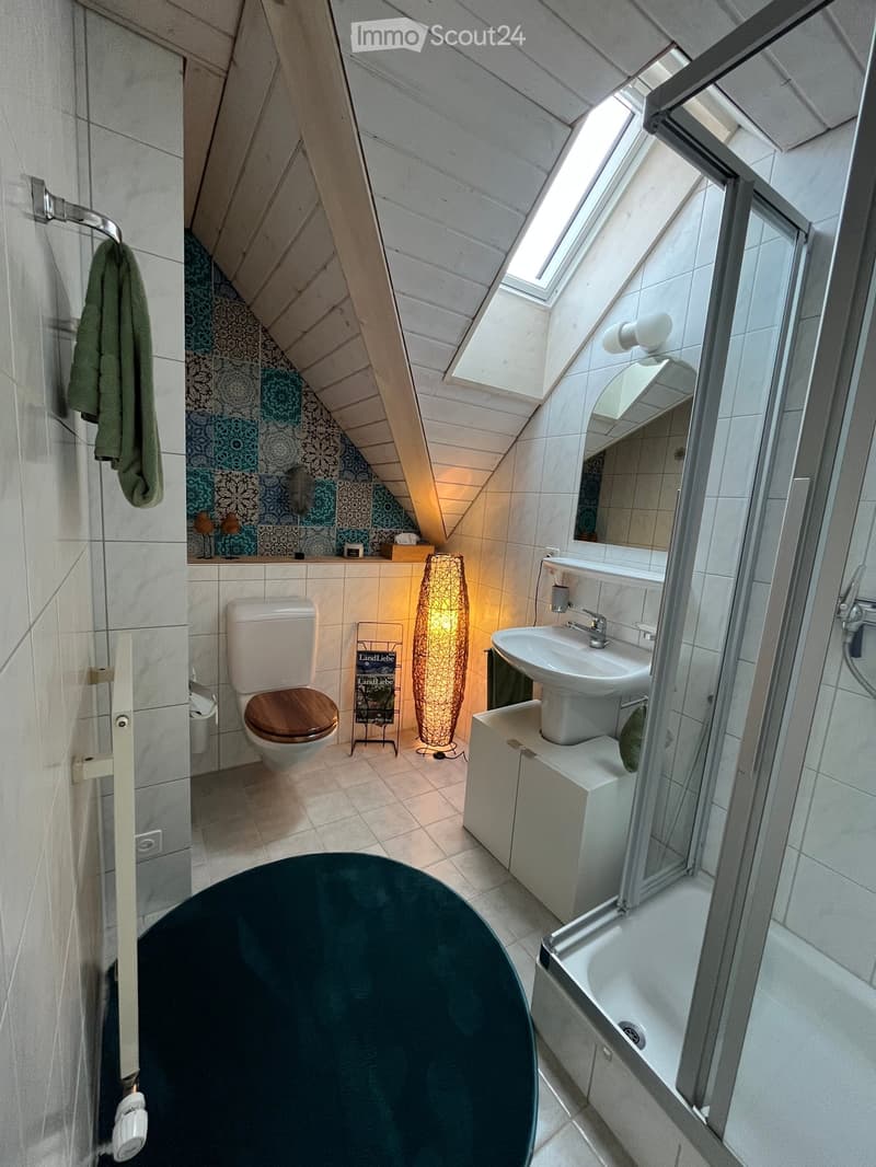 Modern bathroom with a round dark green bathtub, white sink, toilet with wooden seat, and a large window with a skylight above. Features a decorative lamp and a towel hanging on the wall.