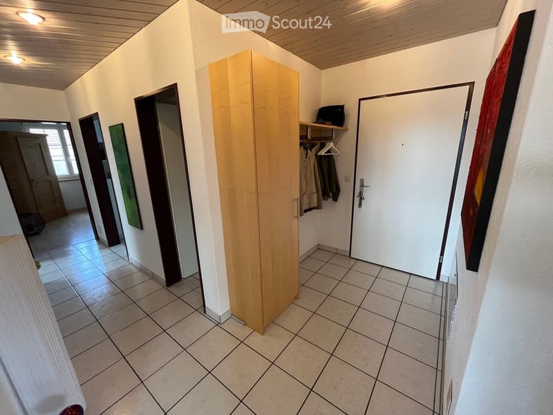 A narrow hallway with tiled flooring, white walls, and a wooden wardrobe. The hallway features several doors and a coat rack with hanging coats.