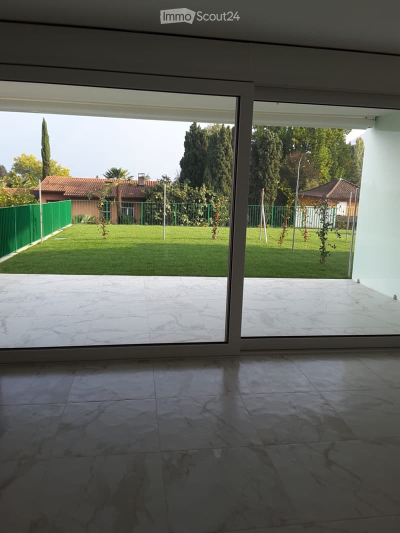 Empty room with large sliding glass doors opening to a well-maintained lawn and garden area. The floor is tiled, and the walls are white.