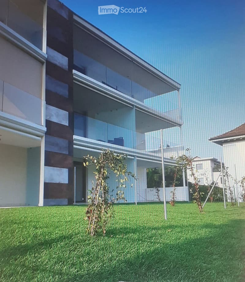 Modern two-story building with large glass windows and balconies, surrounded by a well-maintained lawn and small plants.