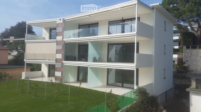 Modern two-story house with white and beige facade, large glass windows, and balconies. The house is surrounded by a well-maintained lawn and small plants.