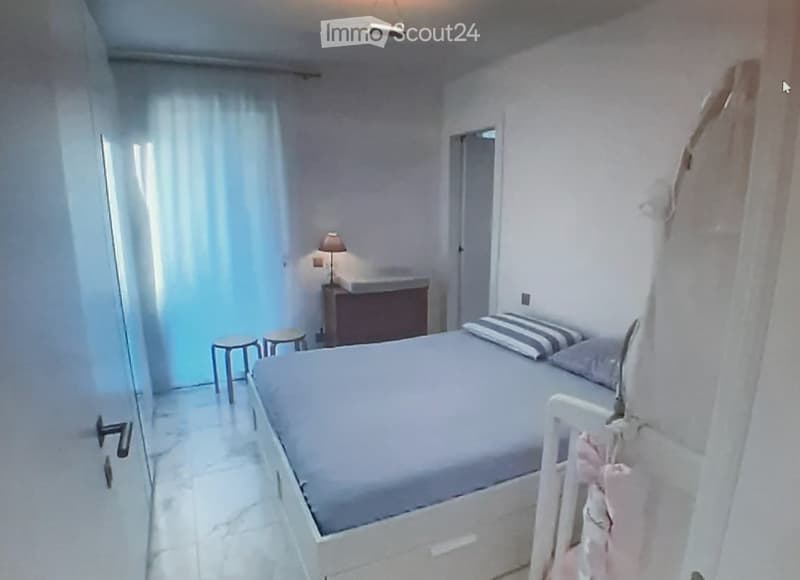 A small, modern bedroom with a single bed, nightstand, lamp, and two stools near the bed. The room features white walls, tiled flooring, and a partially open door leading to a bathroom.