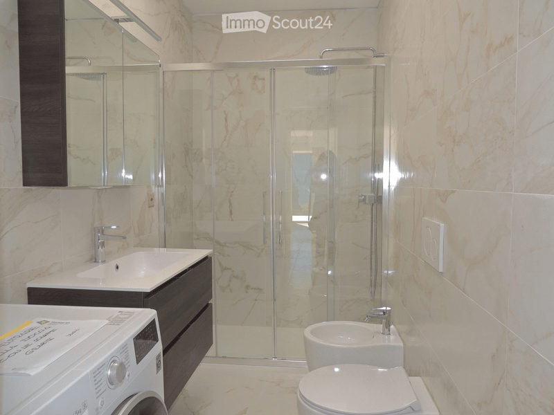 Modern bathroom with marble walls and floors, featuring a glass-enclosed shower, double sinks, a toilet, and a washing machine.
