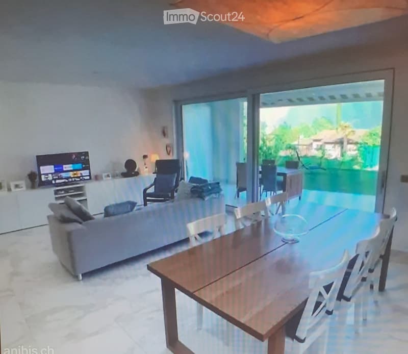 Bright living and dining area with large sliding glass doors leading to a patio, featuring a grey sofa, coffee table, dining table with chairs, and a television on a stand.