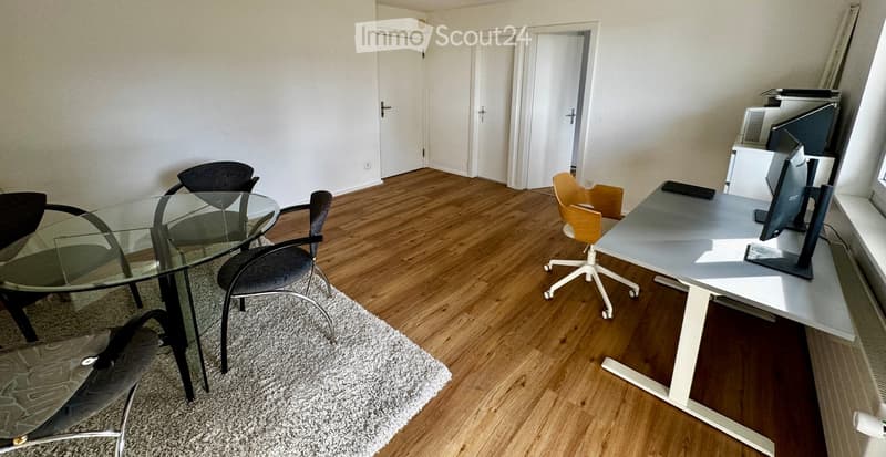Modern office space with wooden flooring, glass table, office chair, desk with computer monitor, and additional chairs.