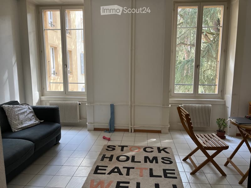 Bright living room with large windows, tiled floor, dark grey sofa, wooden folding chair, small table with laptop, potted plant, and a rug with text "STOCKHOLMS EATLE ME".