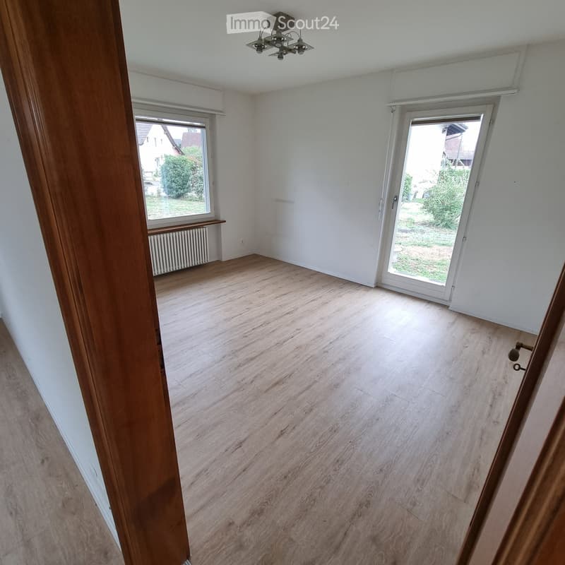 Empty room with light wooden flooring, white walls, large windows, and a sliding glass door leading to a garden area.