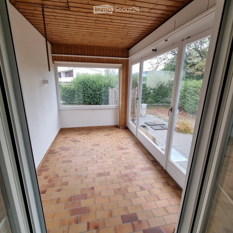 Bright, tiled balcony with wooden ceiling and large windows offering views of greenery and neighboring buildings.