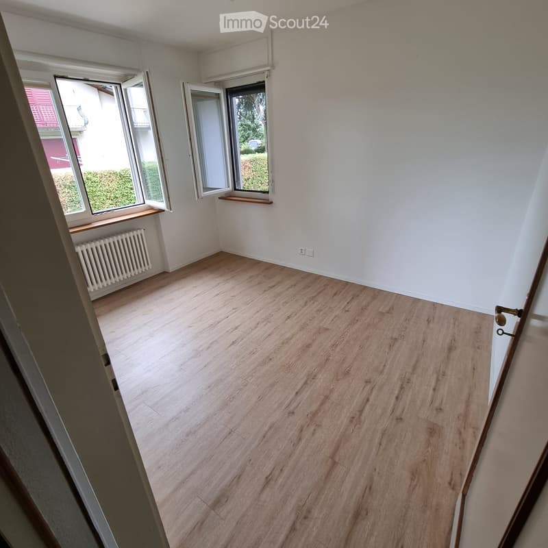 Empty room with light wooden flooring, white walls, and large windows with open blinds. A radiator is visible under one window, and the room has a simple, clean design with no furniture.