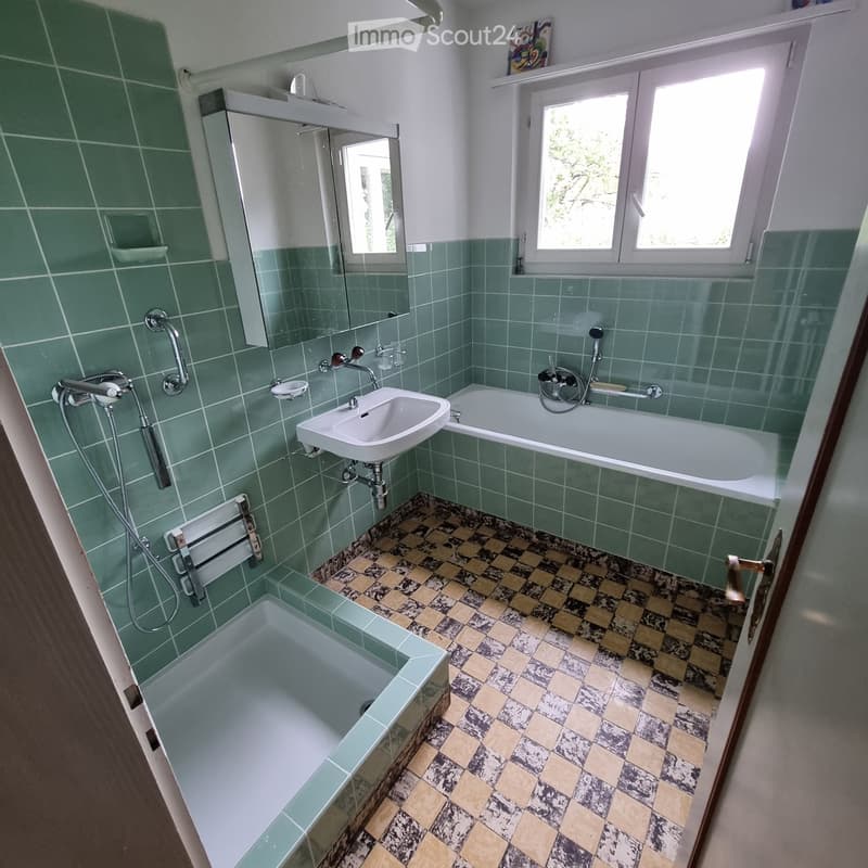 Bathroom with green tiled walls, a white sink, a bathtub, a shower, and a mirror above the sink. The floor has a checkered tile pattern.