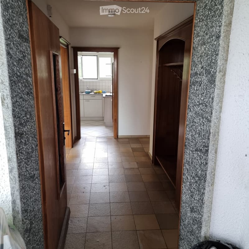 Empty hallway with tiled floor, wooden doors, and a small kitchen visible at the end.