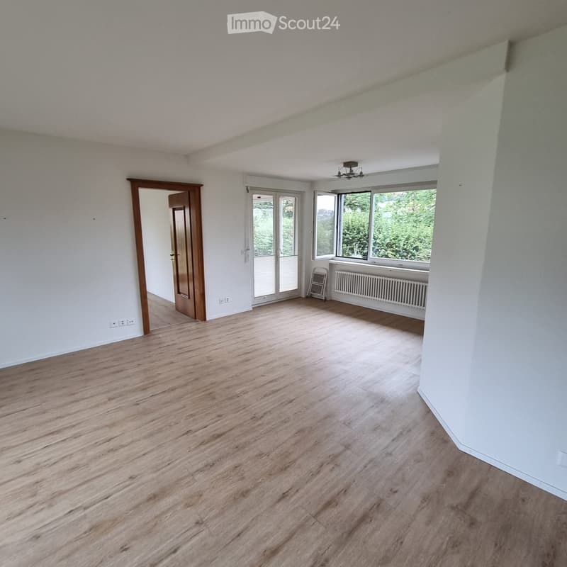 Empty room with light wooden flooring, white walls, large windows, and a sliding glass door leading outside.