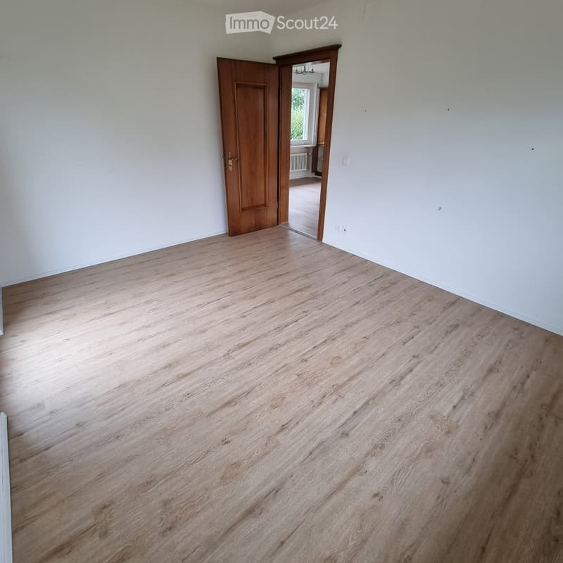 Empty room with light wooden flooring and white walls, featuring an open doorway leading to another room with visible wooden flooring and a window.