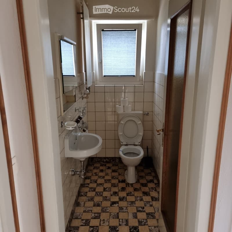 Small bathroom with a toilet, sink, mirror, and toilet paper on a holder. The floor has a checkered tile pattern, and the walls are tiled.