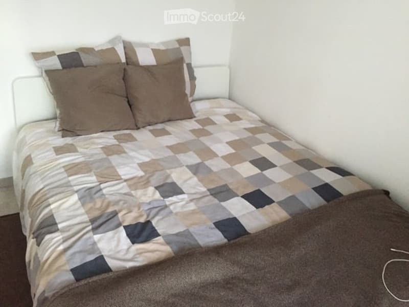 A small bedroom with a single bed, a patterned quilt, and matching pillows. The walls are white, and the floor appears to be wooden.