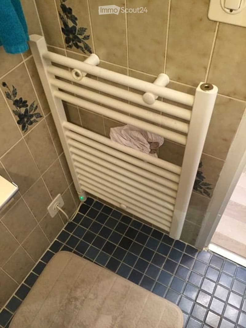 Bathroom with a toilet, a white radiator, and a towel hanging on the wall. The floor is tiled in dark blue, and the walls are tiled in beige with decorative black floral designs.