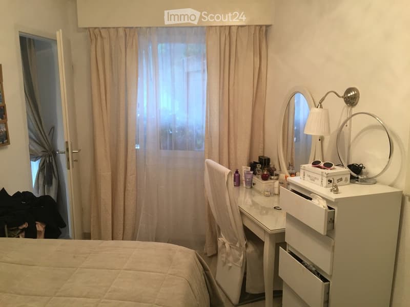 Bedroom with a neatly made bed, a white vanity table with a mirror, a chair, and various cosmetics and personal care items on the vanity.