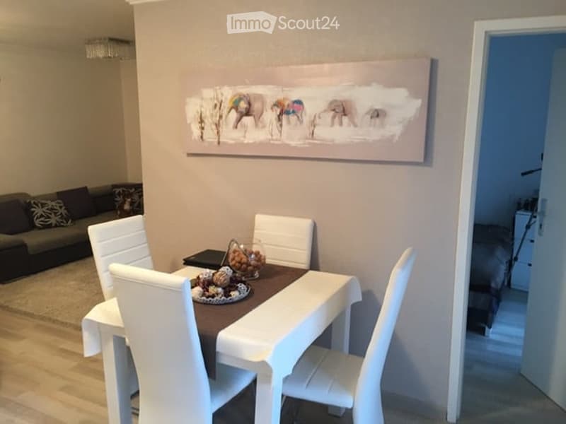 Dining area with a white dining table, four white chairs, a bowl of fruit, and a decorative plate. A grey sofa is visible in the background, along with a large abstract artwork on the wall.