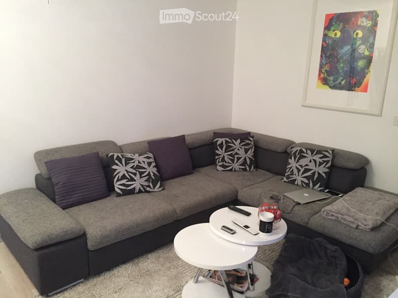 Modern living room with a grey sectional sofa, multiple throw pillows, two white coffee tables, a laptop, and a cup on the coffee table.
