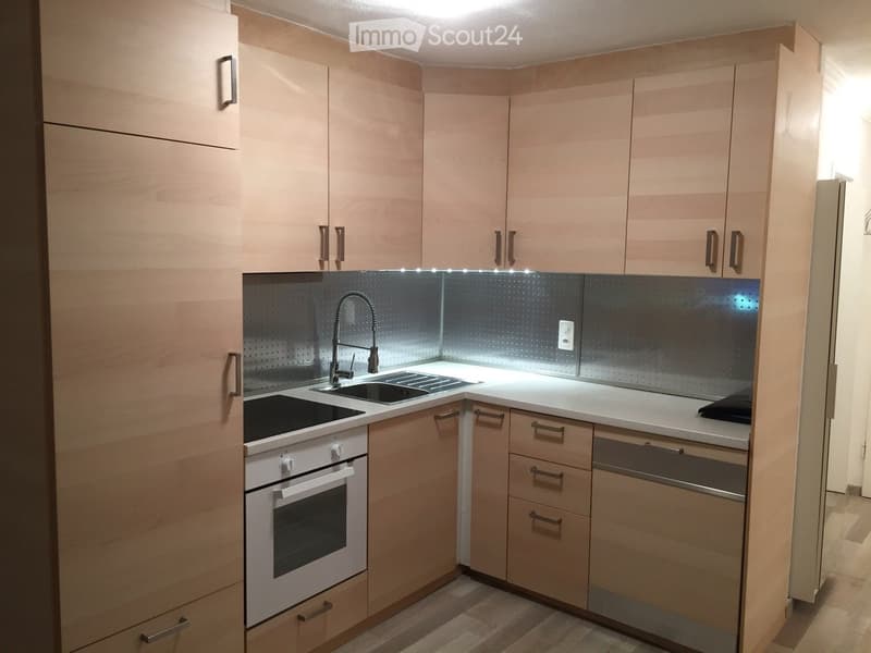 Modern kitchen with light wood cabinets, stainless steel sink, faucet, and built-in oven. Countertop lighting is visible.
