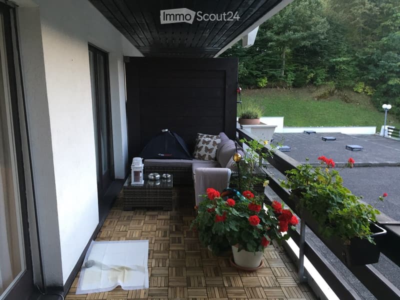Balcony with wooden decking, sofa, small table, potted plants, and a lantern, overlooking a green landscape.