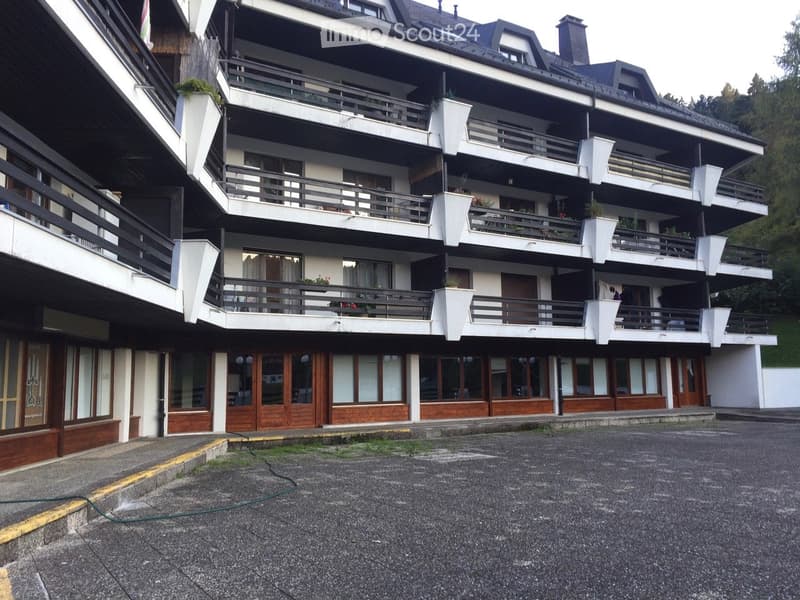 Multi-story building with balconies, large windows, and a covered entrance area. The structure features a mix of white and dark-colored facades with decorative railings and potted plants on the balconies.