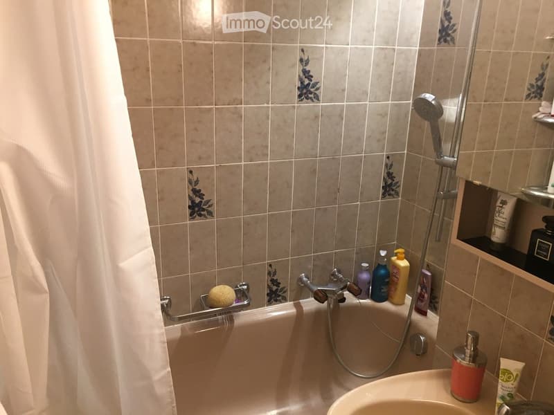 Bathtub with a showerhead and faucet, a white shower curtain, a shelf with various bottles, and a soap dispenser on the sink.