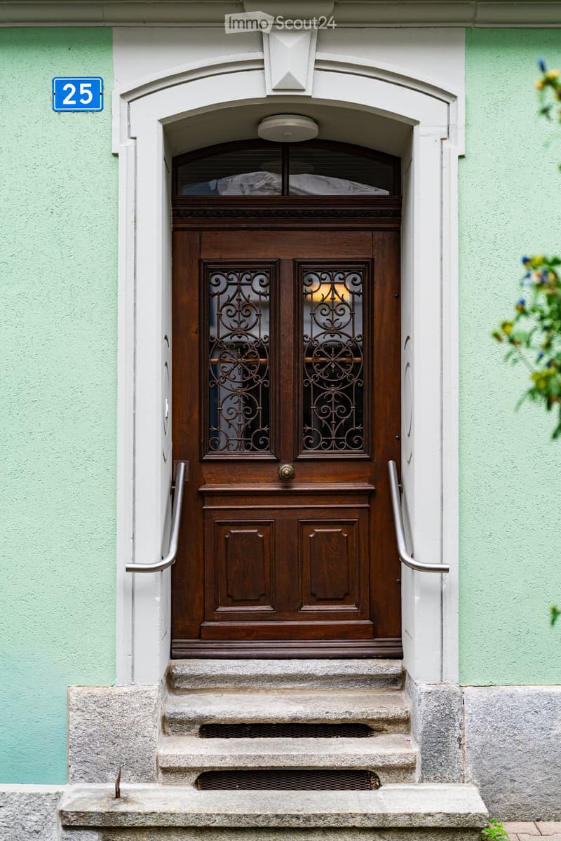 Entrance to a building with a wooden double door featuring ornate glass panels, flanked by light green walls and a small staircase leading up to it.