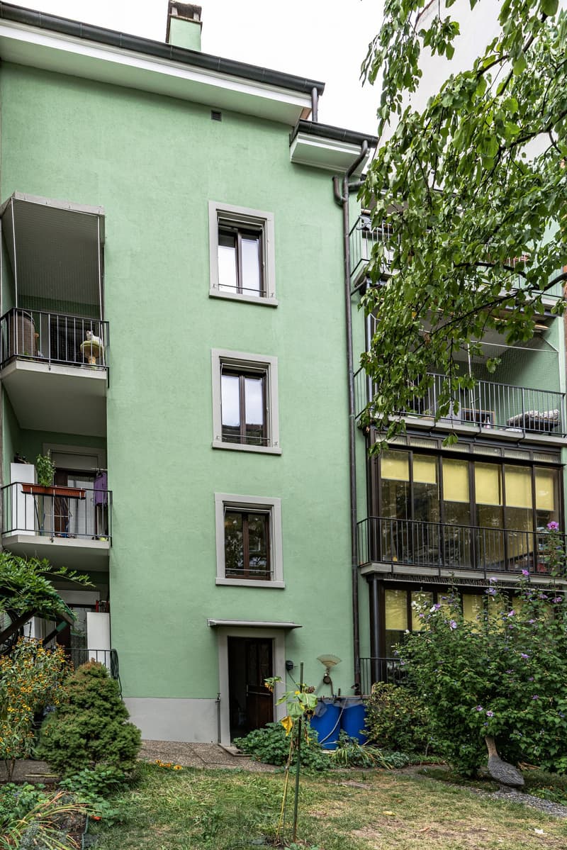 A three-story green apartment building with multiple balconies, large windows, and a small garden area in front.