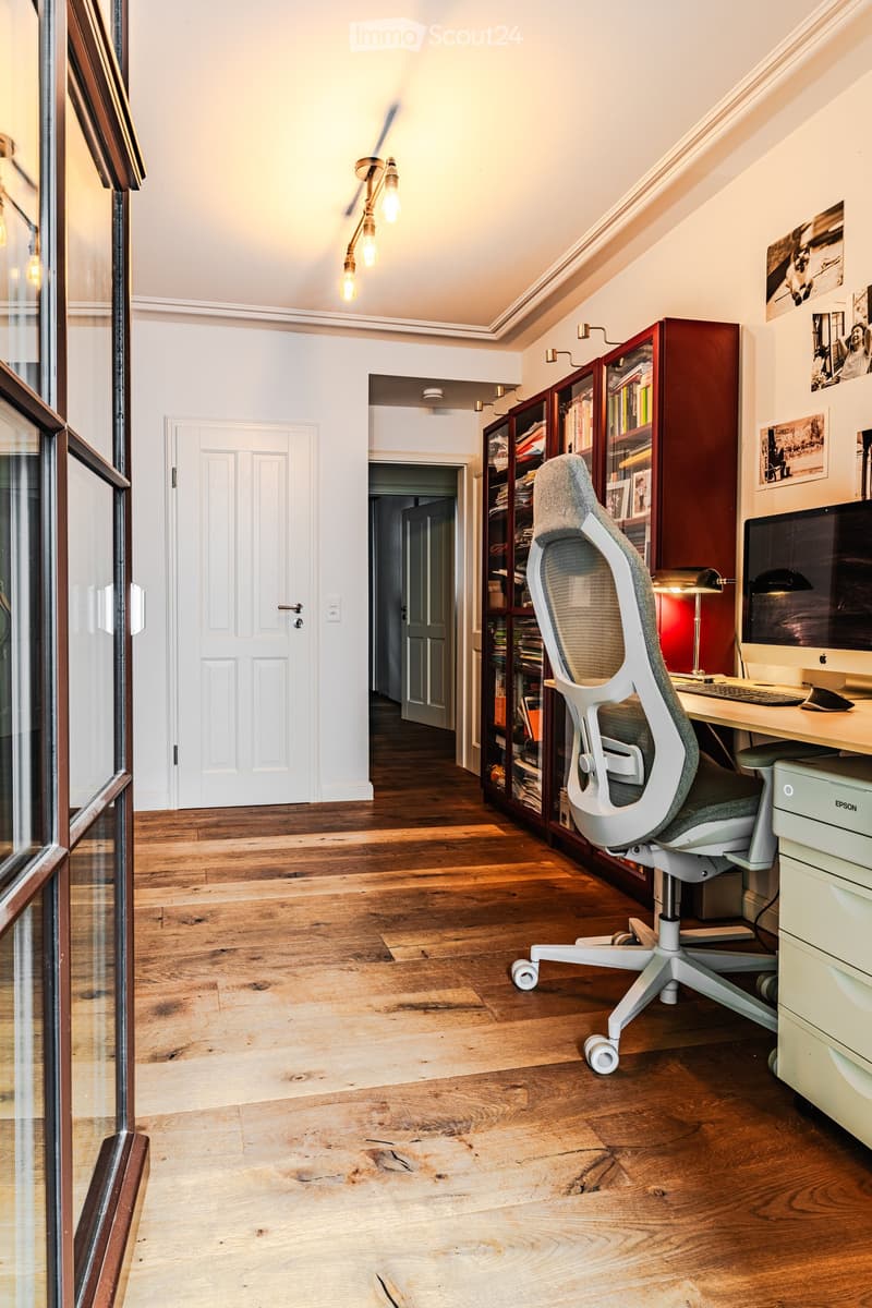 A modern, well-lit study or home office with wooden flooring, large windows, a comfortable office chair, a desk with a computer, and a bookshelf filled with books.