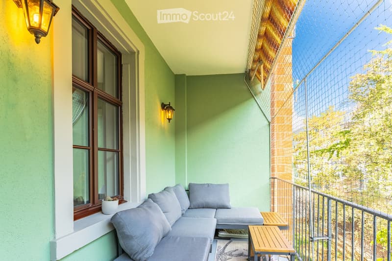 Balcony with green walls, grey sofa with cushions, small wooden tables, potted plant, and a view of trees.